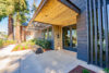 Modern building entrance with covered portico and double glass doors, landscaping features concrete patio, low-lying shrubs, groundcover, and mulch, Contrasting dark-stained wood panels and light-colored concrete