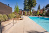 Swimming pool at apartment complex, clear blue water, deck with lounge chairs and seating, two-story buildings with cedar siding, balconies overlooking pool area, tall evergreen trees and landscaping, communal recreational space