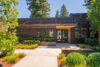 Modern single-story building with wood siding and glass doors Vertical wood slat privacy screen on the right side Concrete walkway with drought-resistant landscaping and planters